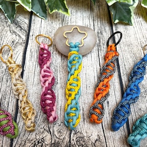 Puede incluir: Una colección de llaveros trenzados coloridos en varios tonos, incluyendo verde, dorado, rosa, amarillo, naranja y azul. Cada llavero tiene un anillo de metal para sujetar llaves. Los llaveros se exhiben sobre una superficie de madera.