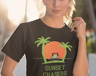 Może przedstawiać: Czarny T-shirt z zielonym i pomarańczowym nadrukiem przedstawiającym jeepa jadącego w stronę zachodu słońca z palmami po obu stronach. Tekst "SUNSET CHASERS" jest nadrukowany na zielono pod grafiką.