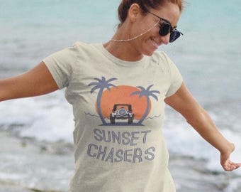 Może przedstawiać: Jasny beżowy t-shirt z grafiką zachodu słońca, palm i jeepa. Tekst "Sunset Chasers" jest wydrukowany poniżej w kolorze niebieskim. Koszulka ma krótkie rękawy.