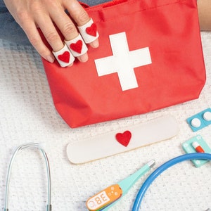 May include: A red first aid kit with a white cross, a stethoscope, a thermometer, a bandage, and a small box of pills. The kit is on a white surface.