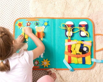 May include: A blue and orange felt activity book with various textures and activities for children. The book features a bear, a flower, shoes, and a pillow with buckles and straps.