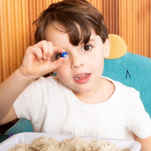 May include: A young child holds a small, blue and clear, multi-sided gem up to their eye. The child is wearing a white t-shirt and sitting in a teal chair. A tray of sand is in the foreground.