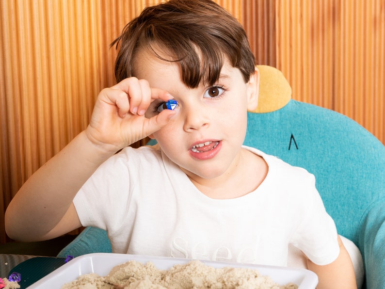 May include: A young child holds a small, blue and clear, multi-sided gem up to their eye. The child is wearing a white t-shirt and sitting in a teal chair. A tray of sand is in the foreground.