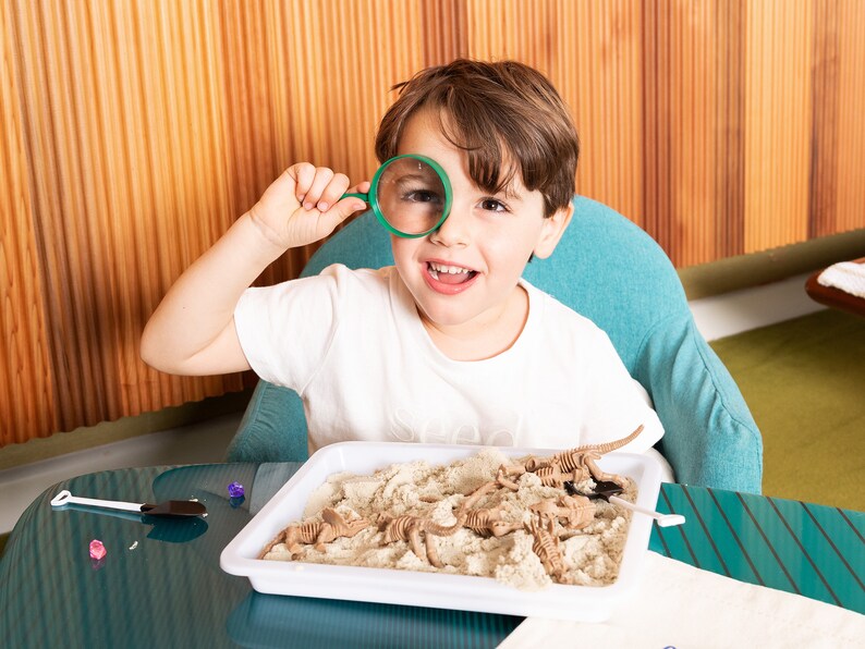 May include: A young child with brown hair and a white t-shirt looks through a green magnifying glass. A white tray on a teal table contains sand and brown dinosaur skeleton toys. A small white shovel is also on the table.