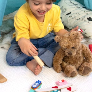 May include: A child plays doctor with a teddy bear. The child is holding a wooden syringe and the teddy bear has a bandage on its ear. There are toy medical supplies on the floor, including a thermometer, bandages, and pills.