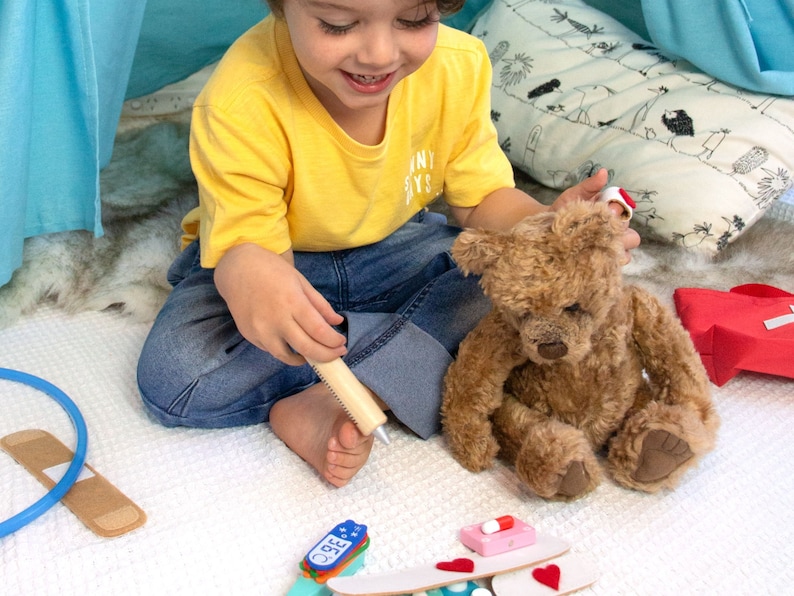 May include: A child plays doctor with a teddy bear. The child is holding a wooden syringe and the teddy bear has a bandage on its ear. There are toy medical supplies on the floor, including a thermometer, bandages, and pills.