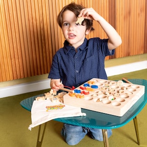 K&ouml;nnte beinhalten: Ein Kind spielt mit einem Angelspiel aus Holz. Das Spiel besteht aus einem Holzbrett mit L&ouml;chern, bunten Formen und kleinen Holzfischen. Ein t&uuml;rkisfarbener Tisch tr&auml;gt das Spiel, und eine wei&szlig;e Tasche mit "Axel Adventures" ist ebenfalls zu sehen.