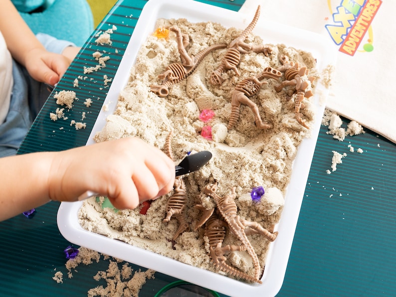 May include: A white plastic tray filled with tan sand and several brown plastic dinosaur skeletons. Colorful gems are scattered throughout the sand. A child's hand is using a black scoop to dig in the sand. A canvas bag with the text "Axx Adventures" is in the background.