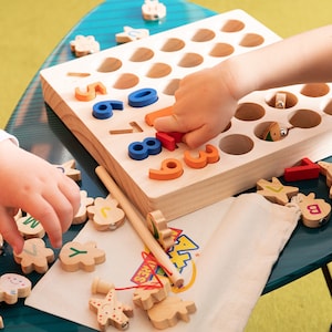 K&ouml;nnte beinhalten: Holz-Lernspielzeug mit bunten Zahlen und Buchstabenformen. Das Set enth&auml;lt eine Holztafel mit L&ouml;chern, einen kleinen Beutel und verschiedene Holzteile. Kinderh&auml;nde interagieren mit dem Spielzeug auf einem blauen Tisch.