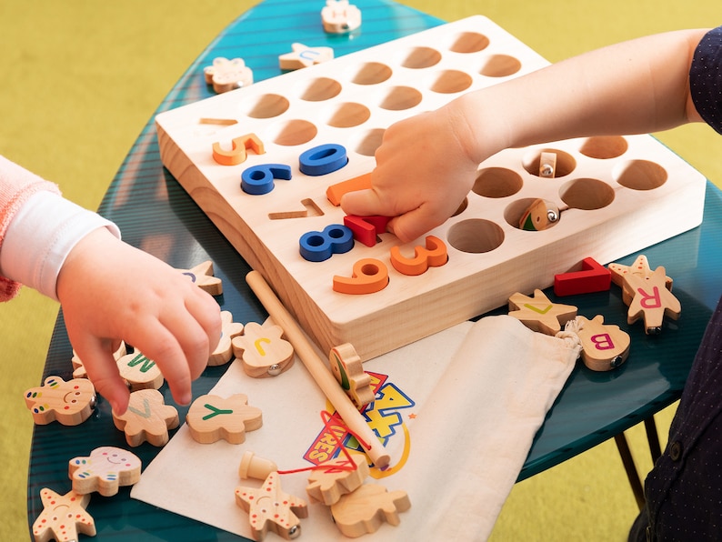 K&ouml;nnte beinhalten: Holz-Lernspielzeug mit bunten Zahlen und Buchstabenformen. Das Set enth&auml;lt eine Holztafel mit L&ouml;chern, einen kleinen Beutel und verschiedene Holzteile. Kinderh&auml;nde interagieren mit dem Spielzeug auf einem blauen Tisch.