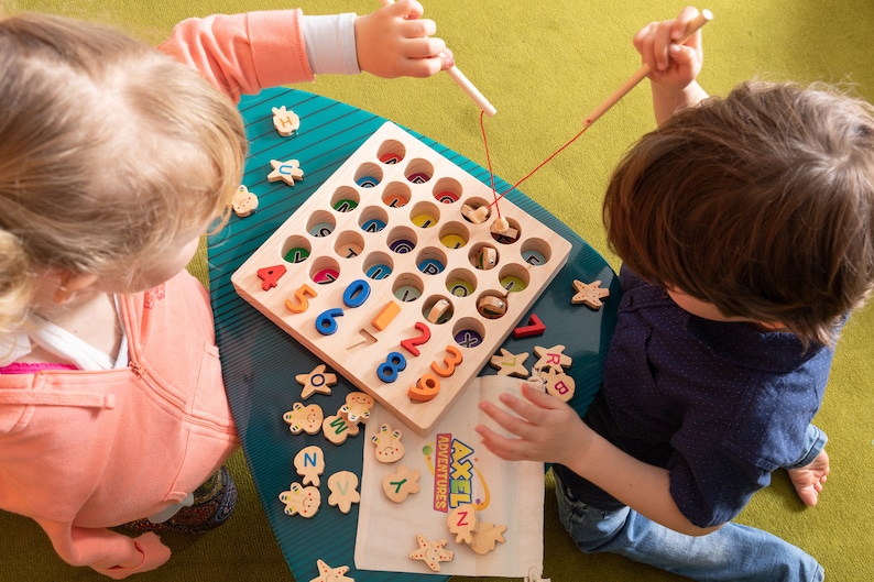 K&ouml;nnte beinhalten: Holz-Lernspielzeug mit bunten Zahlen und Buchstaben. Kinder spielen mit einem Holz-Angelspiel, bei dem sie mit einer Magnetangel Holzformen fischen. Das Spiel enth&auml;lt ein Holzbrett mit L&ouml;chern und einen Stoffbeutel mit dem Text "AXEL ADVENTURES".