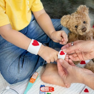 May include: A child wearing a blue jean outfit and a yellow shirt is playing doctor with a teddy bear. The child is wearing a white armband with a red heart on it. The child is also wearing a toy stethoscope around their neck. The child is holding a toy thermometer in their hand. The teddy bear is wearing a white armband with a red heart on it.