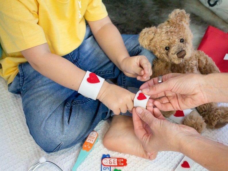 May include: A child wearing a blue jean outfit and a yellow shirt is playing doctor with a teddy bear. The child is wearing a white armband with a red heart on it. The child is also wearing a toy stethoscope around their neck. The child is holding a toy thermometer in their hand. The teddy bear is wearing a white armband with a red heart on it.