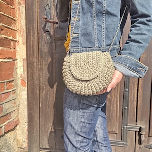 Pu&ograve; includere: Una persona che indossa una giacca di jeans e jeans porta una borsa a tracolla in crochet beige con una catena argentata.