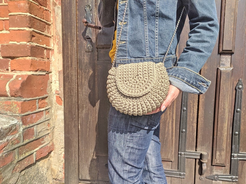 Pu&ograve; includere: Una persona che indossa una giacca di jeans e jeans porta una borsa a tracolla in crochet beige con una catena argentata.