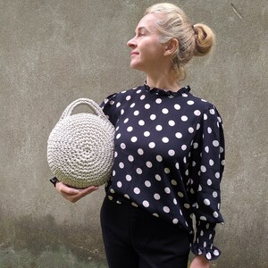 May include: A woman is holding a round, beige crocheted bag. She is wearing a black and white polka dot blouse and black pants.