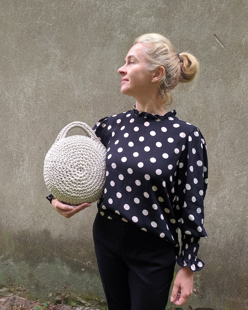 May include: A woman is holding a round, beige crocheted bag. She is wearing a black and white polka dot blouse and black pants.
