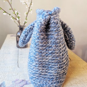 May include: A blue and white knitted bag with two small handles. The bag is made of thick yarn and has a textured appearance. The bag is sitting on a distressed white wooden surface. A vase with white flowers is in the background.