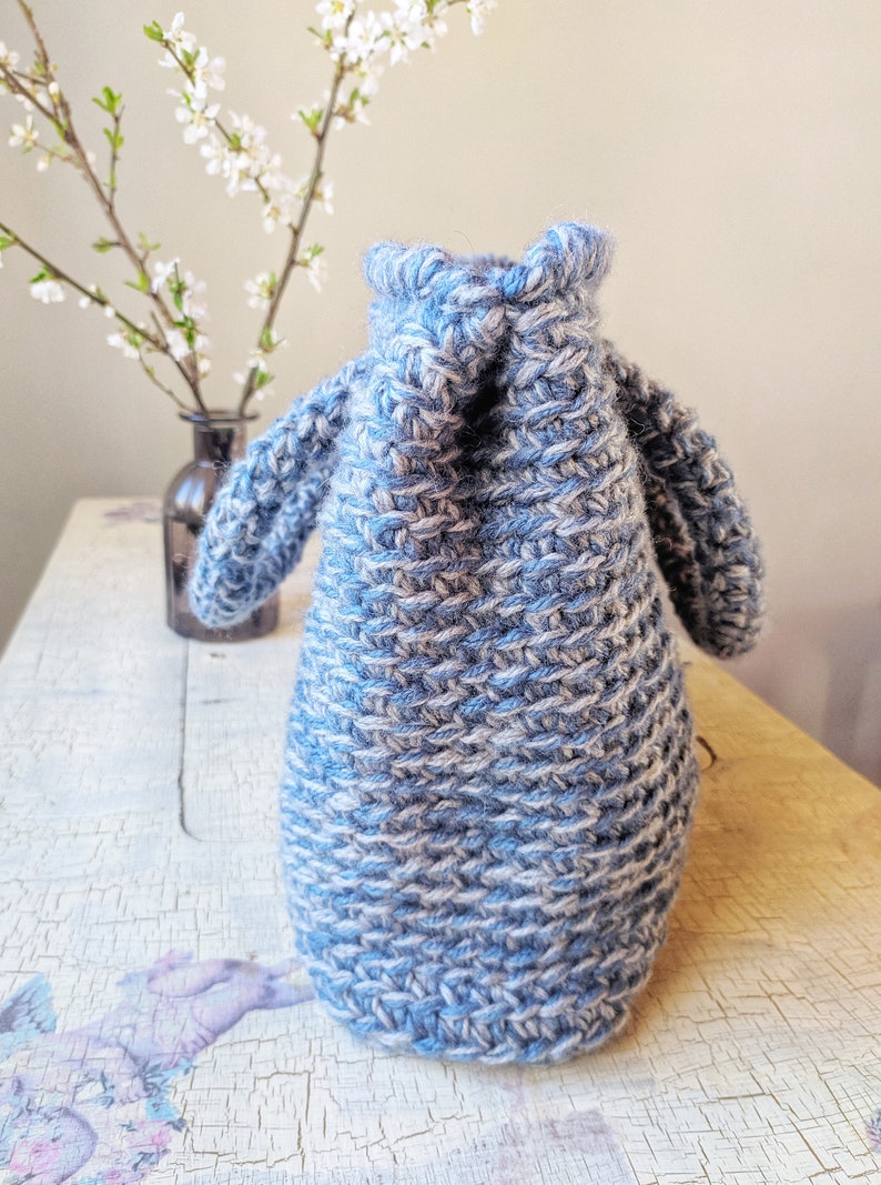 May include: A blue and white knitted bag with two small handles. The bag is made of thick yarn and has a textured appearance. The bag is sitting on a distressed white wooden surface. A vase with white flowers is in the background.