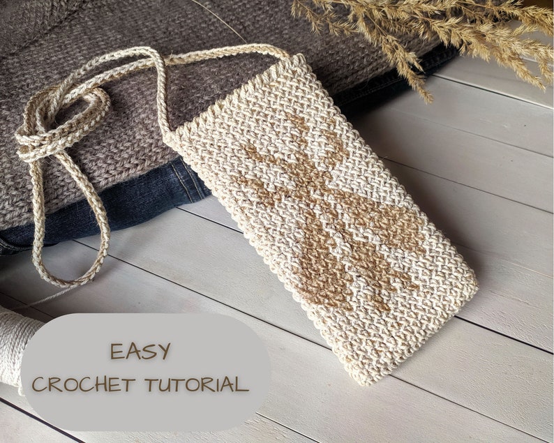 May include: A close-up of a crocheted beige and white crossbody bag with a long strap. The bag is lying on a white wooden surface. The text "EASY CROCHET TUTORIAL" is printed on a gray oval label below the bag.