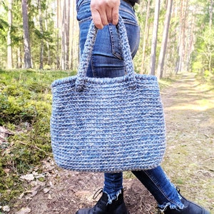 May include: A blue and white crocheted tote bag with sturdy handles. The bag is made of a thick, textured yarn and is being held outdoors. The bag's color is a mix of light and dark blue, creating a striped effect.