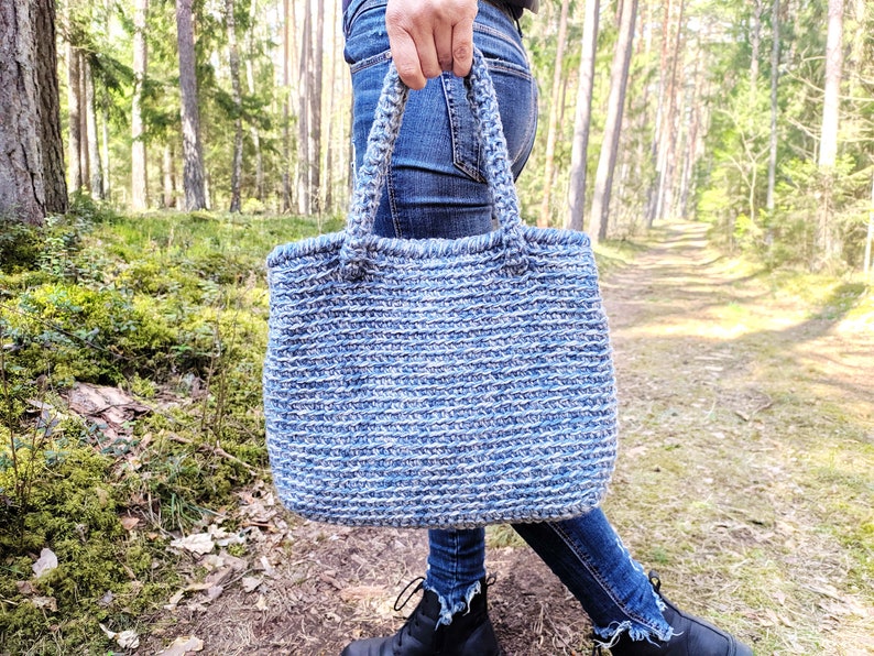 May include: A blue and white crocheted tote bag with sturdy handles. The bag is made of a thick, textured yarn and is being held outdoors. The bag's color is a mix of light and dark blue, creating a striped effect.