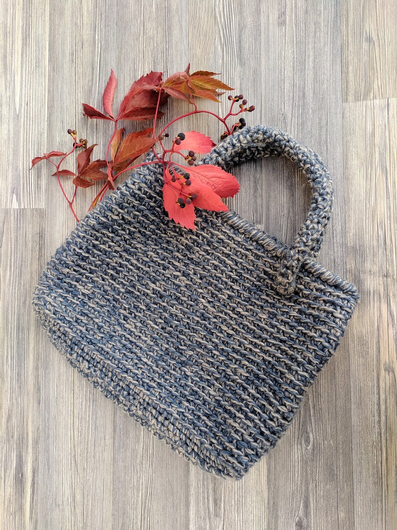 May include: A handmade, blue and beige crocheted tote bag with a sturdy handle. The bag is rectangular and has a textured appearance. Red and orange fall leaves and berries are placed on the bag and wooden surface.