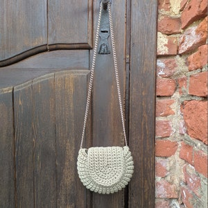 Pu&ograve; includere: Una borsa a mano di colore grigio chiaro realizzata a uncinetto a forma di mezzaluna con una tracolla a catena argentata appesa a una porta di legno.