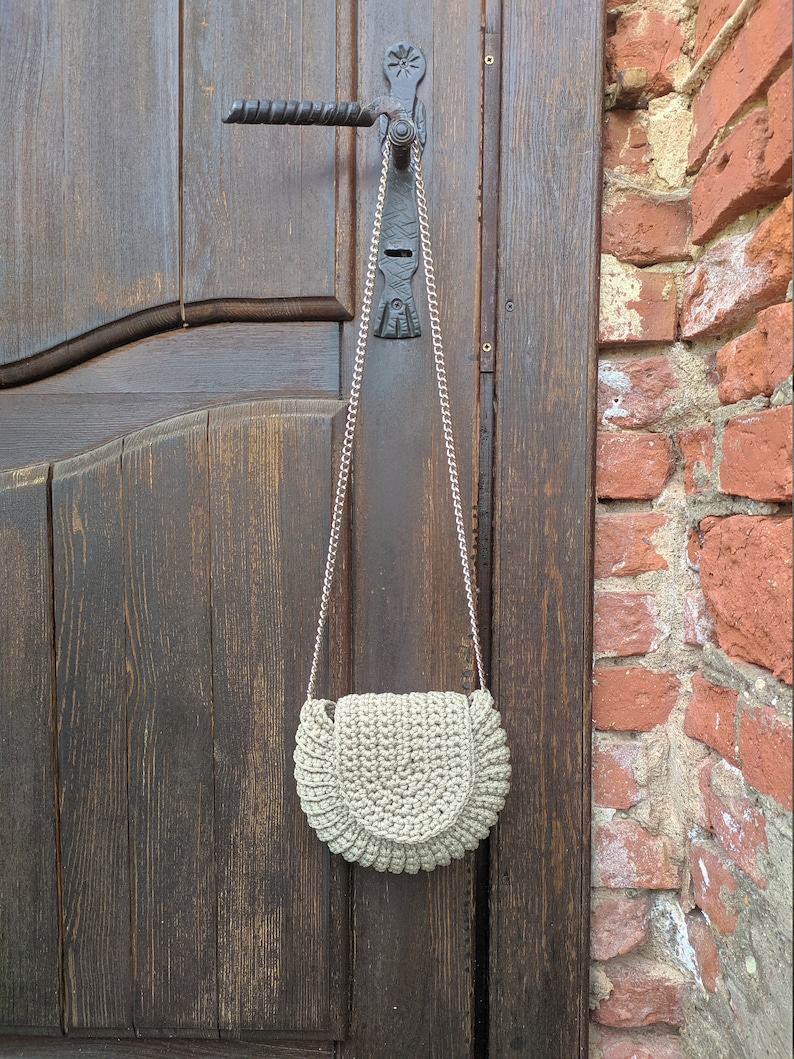 Pu&ograve; includere: Una borsa a mano di colore grigio chiaro realizzata a uncinetto a forma di mezzaluna con una tracolla a catena argentata appesa a una porta di legno.