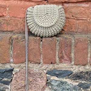 Pu&ograve; includere: Una borsa in crochet beige con una tracolla a catena argentata. La borsa &egrave; appesa a un muro di mattoni.