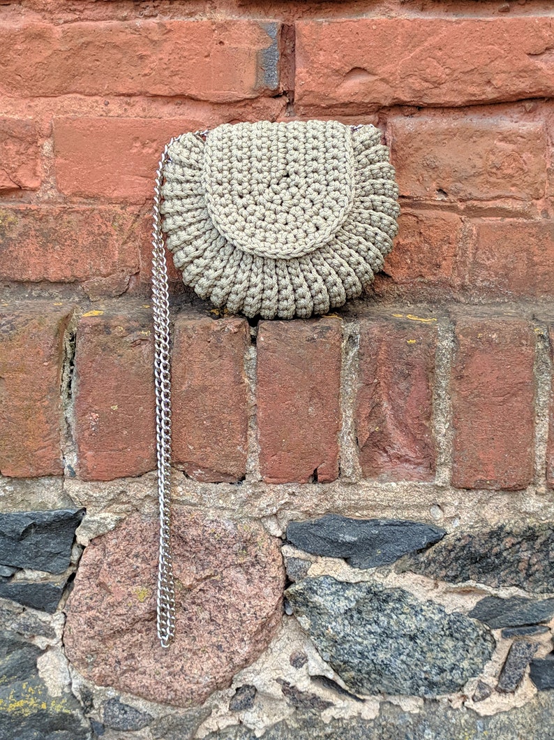 Pu&ograve; includere: Una borsa in crochet beige con una tracolla a catena argentata. La borsa &egrave; appesa a un muro di mattoni.