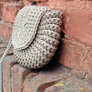 Pu&ograve; includere: Una borsa in crochet beige con chiusura a lembo e una tracolla a catena argentata. La borsa &egrave; appoggiata a un muro di mattoni.