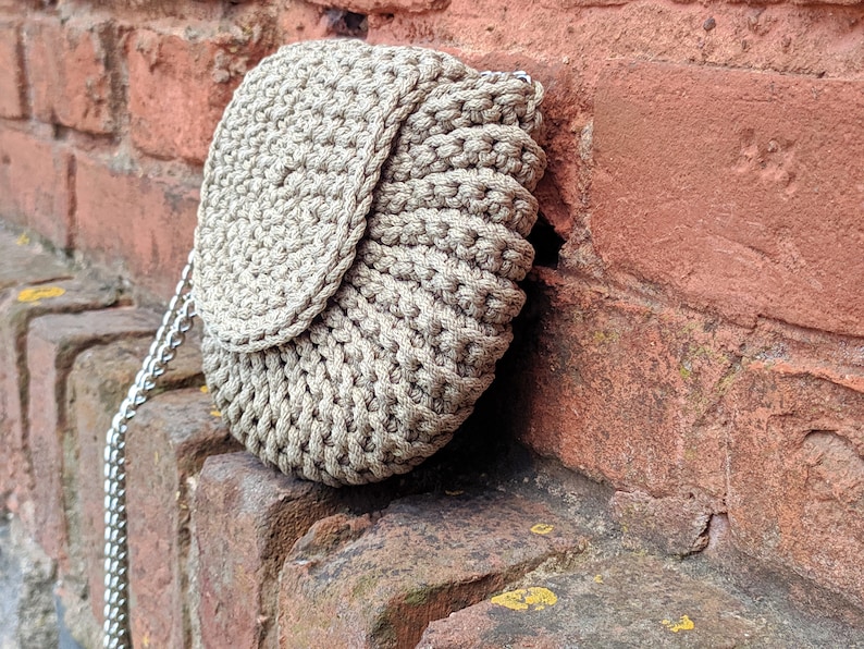 Pu&ograve; includere: Una borsa in crochet beige con chiusura a lembo e una tracolla a catena argentata. La borsa &egrave; appoggiata a un muro di mattoni.
