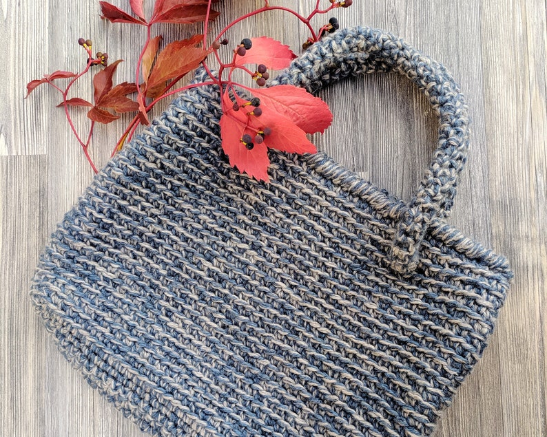 May include: A handmade, blue and beige crocheted handbag with a sturdy handle. The bag has a rectangular shape and is decorated with red leaves and berries. The bag is made of a thick yarn, giving it a textured appearance.