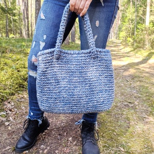 May include: A handmade blue and white crochet tote bag with sturdy handles. The bag is held in front of a person wearing blue jeans and black boots. The bag's textured design is visible, showcasing the craftsmanship.