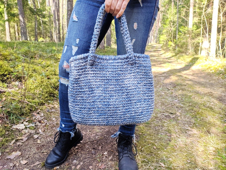 May include: A handmade blue and white crochet tote bag with sturdy handles. The bag is held in front of a person wearing blue jeans and black boots. The bag's textured design is visible, showcasing the craftsmanship.