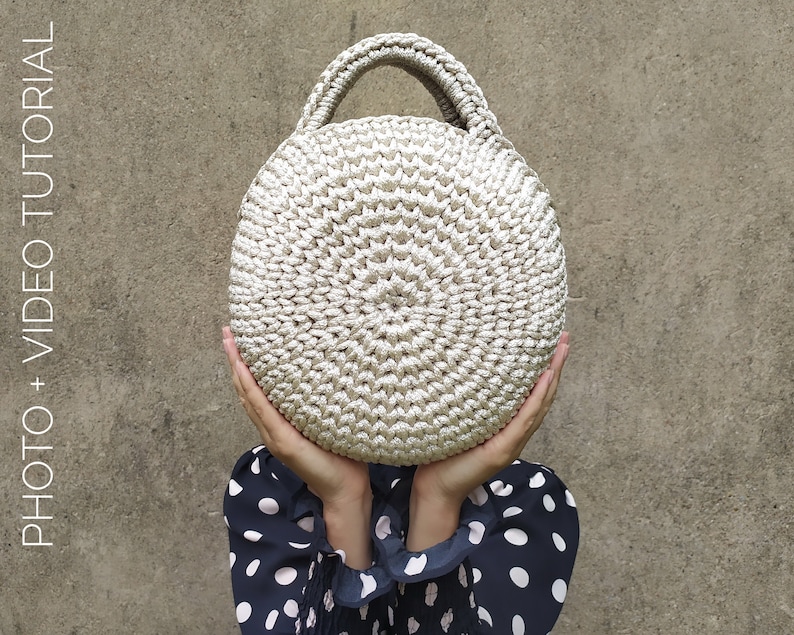 May include: A round crocheted bag with a beige color and a single handle. The bag is held in front of a person wearing a black and white polka dot shirt.