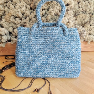 May include: A handmade blue and white knitted tote bag with two handles. The bag has a rectangular shape and a textured appearance. A long, dark brown strap with wooden beads is lying next to the bag.