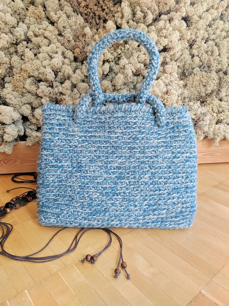 May include: A handmade blue and white knitted tote bag with two handles. The bag has a rectangular shape and a textured appearance. A long, dark brown strap with wooden beads is lying next to the bag.