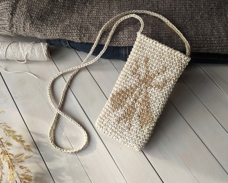 May include: A small crocheted bag with a beige and brown geometric pattern. The bag has a long strap and is made of natural fibers.