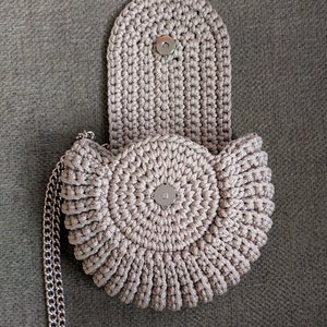 Pu&ograve; includere: Una borsa a mano in crochet beige di forma circolare con chiusura a lembo. La borsa ha una tracolla a catena argentata.