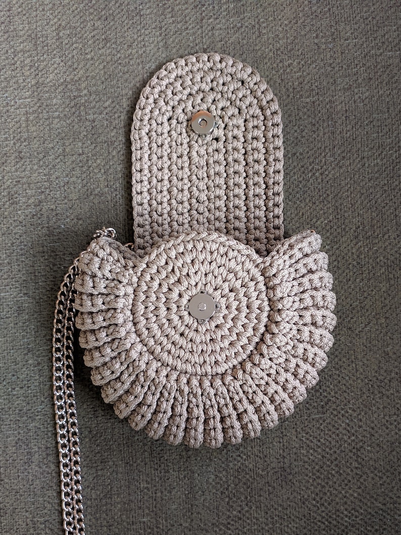 Pu&ograve; includere: Una borsa a mano in crochet beige di forma circolare con chiusura a lembo. La borsa ha una tracolla a catena argentata.