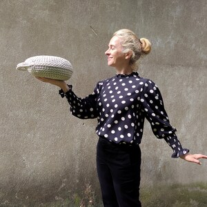 May include: A woman in a black and white polka dot blouse holds a white crocheted oval-shaped basket in front of a gray wall.