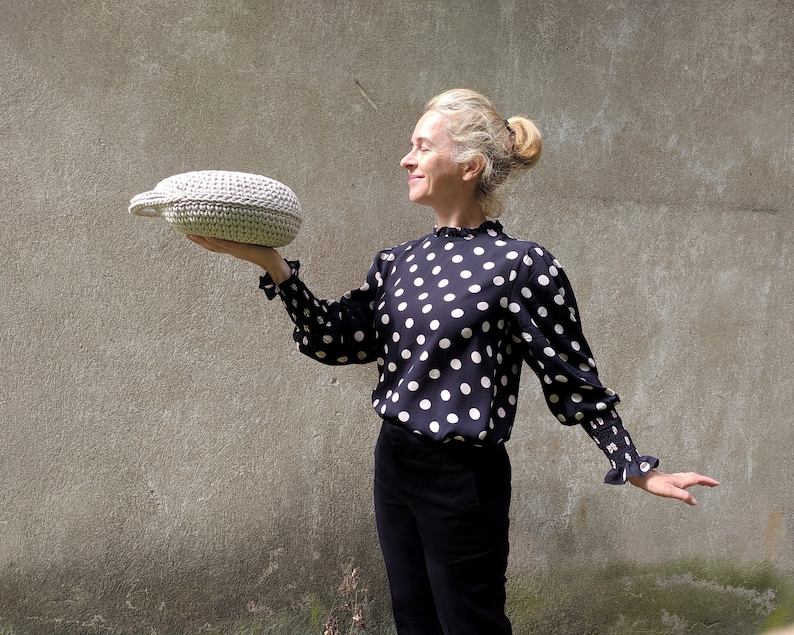 May include: A woman in a black and white polka dot blouse holds a white crocheted oval-shaped basket in front of a gray wall.