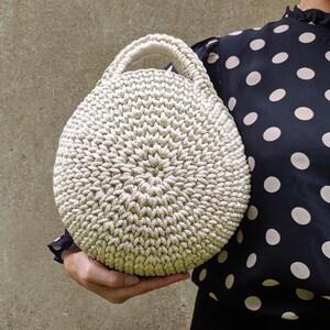 May include: A round, crocheted handbag in a cream color. The bag has a single handle and a textured, woven design.