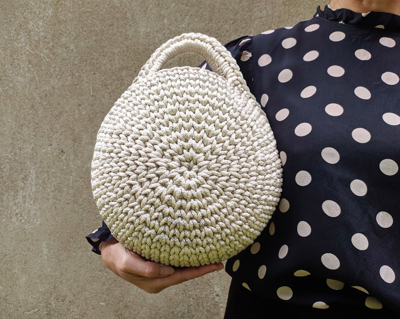 May include: A round, crocheted handbag in a cream color. The bag has a single handle and a textured, woven design.