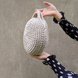 May include: A round, crocheted bag with a cream-colored design. The bag has a single handle and is held by a hand.