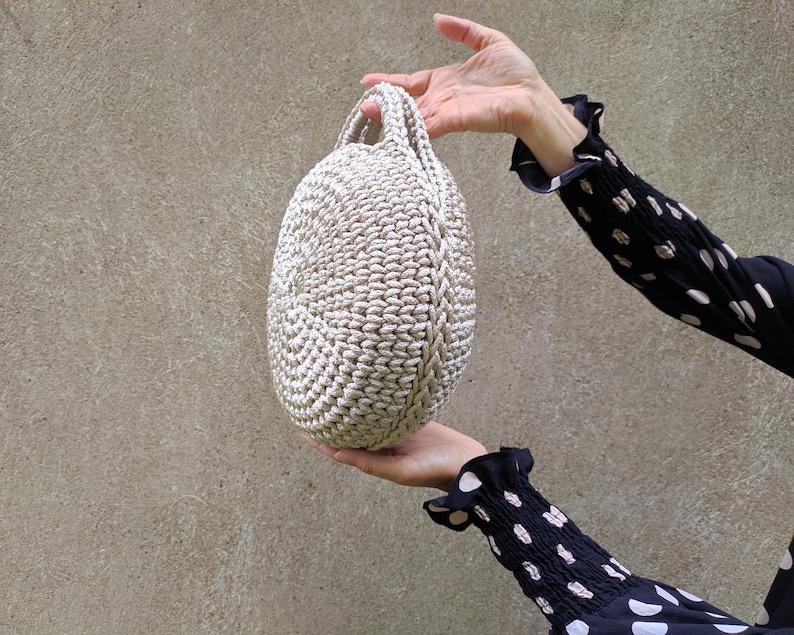 May include: A round, crocheted bag with a cream-colored design. The bag has a single handle and is held by a hand.