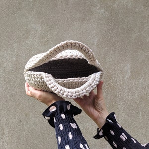 May include: A beige crocheted tote bag with a wide opening, held by two hands.