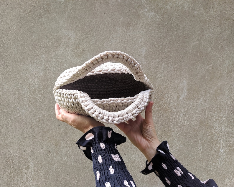 May include: A beige crocheted tote bag with a wide opening, held by two hands.